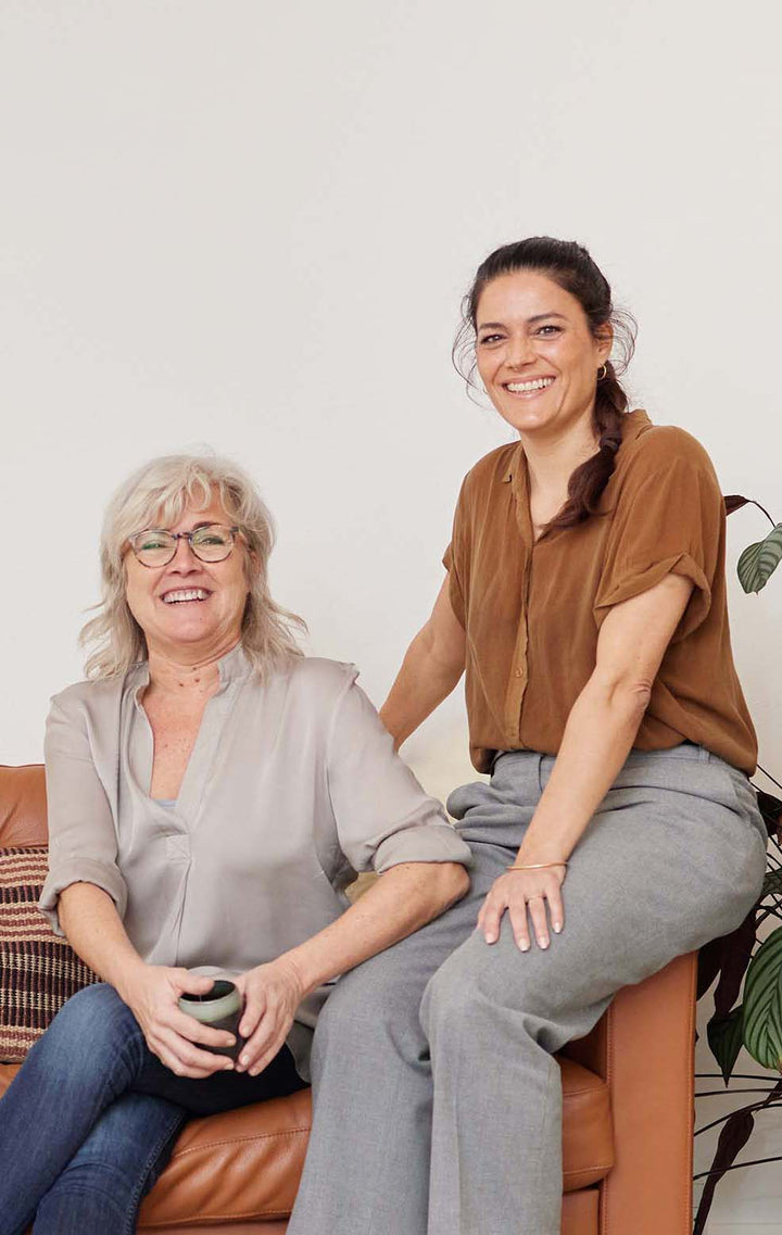 Camilla og Tine der ejer Care by Nature sidder sammen og smiler i brun lædersofa