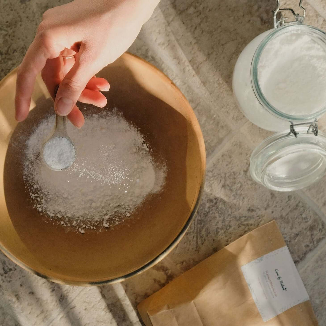 Natriumbikarbonat drysses i keramikskål til naturlig rengøring fra et glas med bikarbonat ved siden af en papirspose og med stenbaggrund